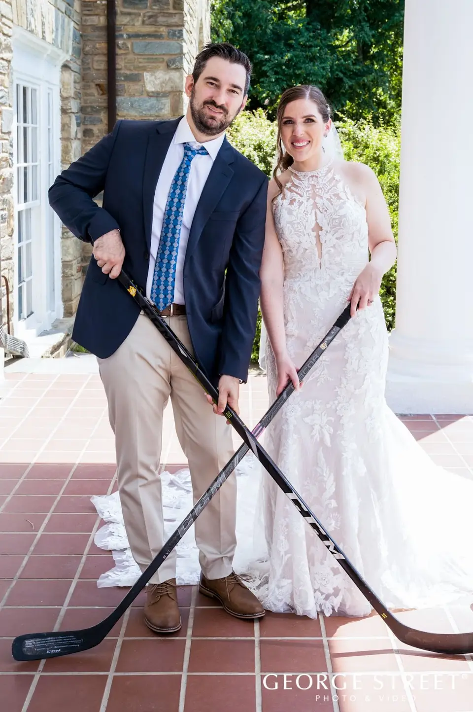 Bride and groom posing on a brick patio with crossed hockey sticks in front of a white-columned building