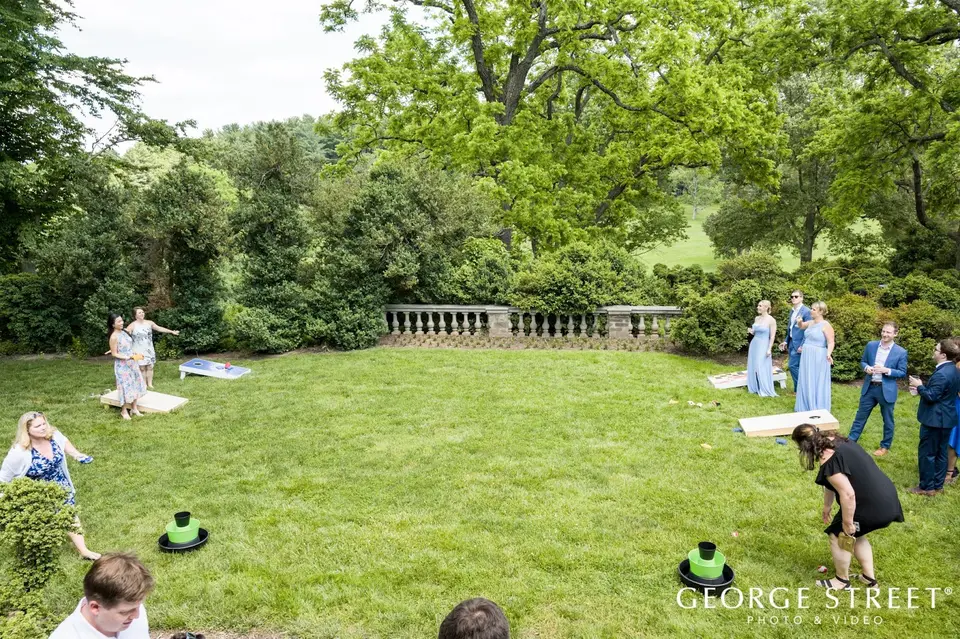 Wedding guests playing cornhole on a large lawn with a stone balustrade and trees behind them