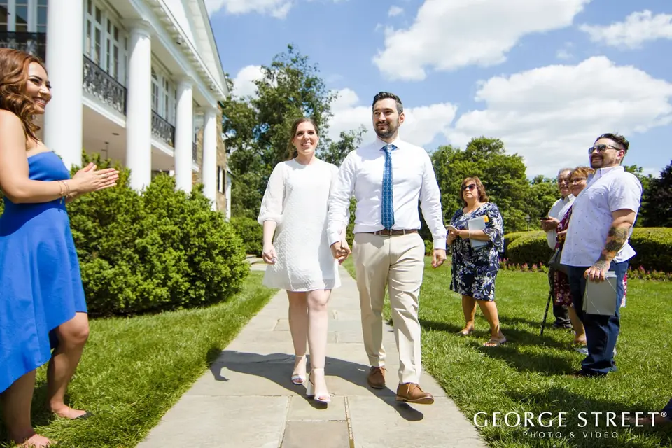 Bride in a short white dress and groom in a blue tie walking a garden path as guests watch