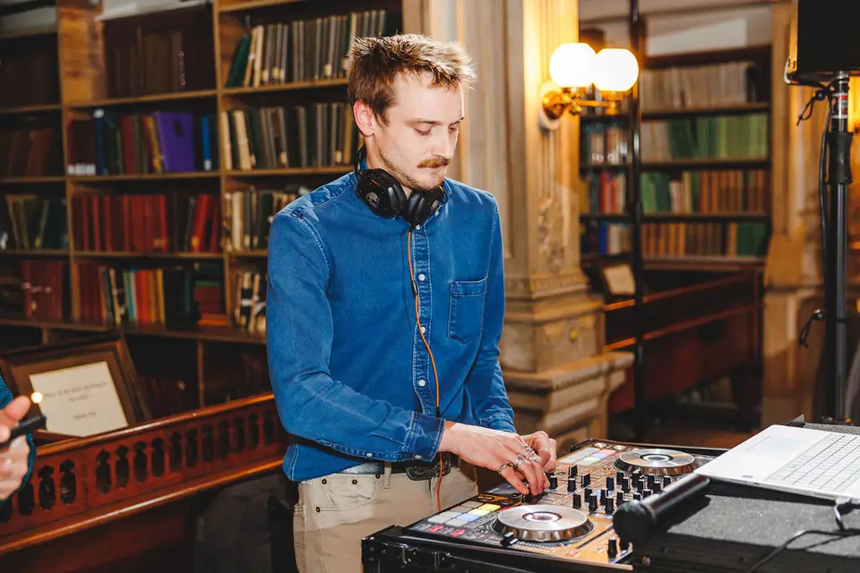 DJ in a denim shirt and headphones working at a DJ controller in a library with bookshelves