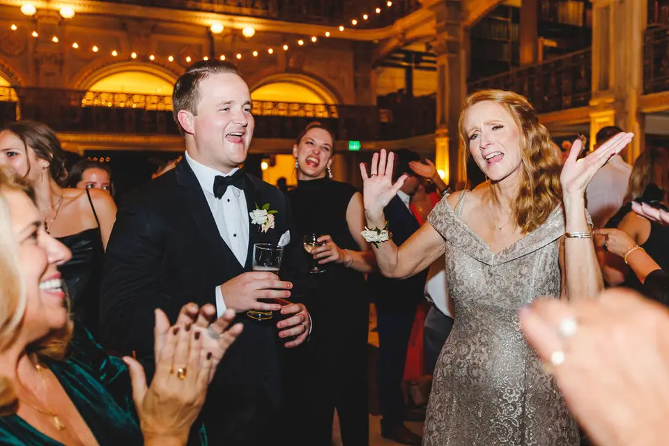Man in a tuxedo singing and a woman in a gold dress dancing with raised hands among clapping guests