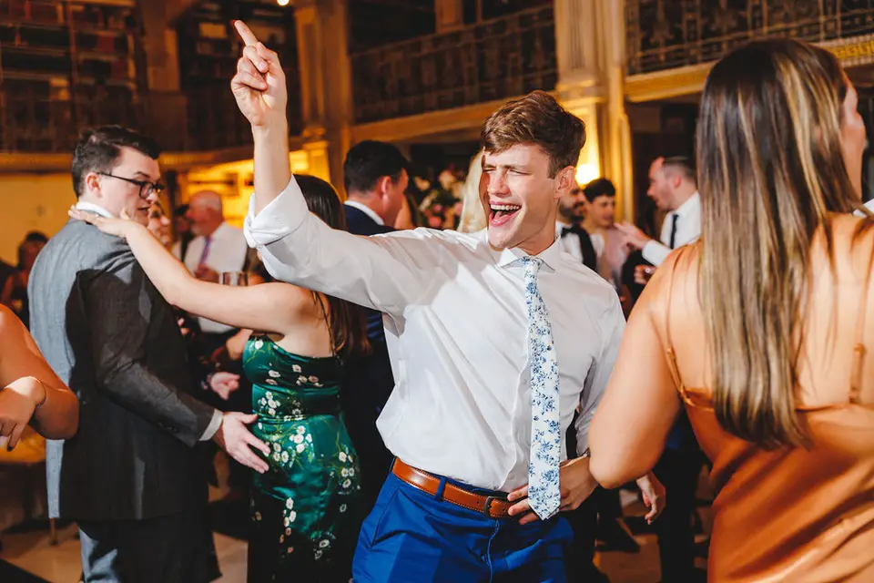 Man in a white shirt and floral tie laughing and pointing upward while dancing at a reception