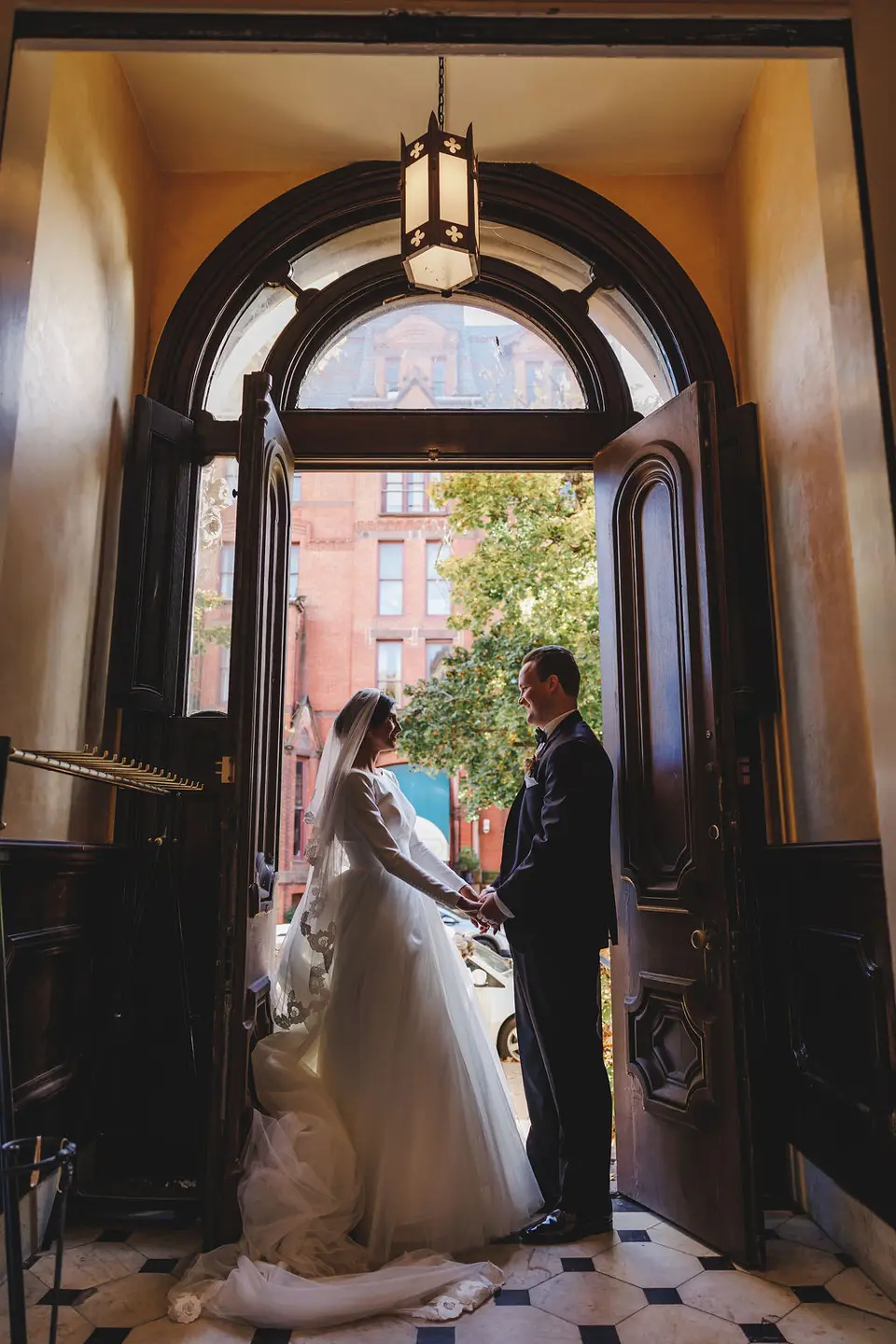 Bride and groom holding hands in a large arched wooden doorway with a brick building outside