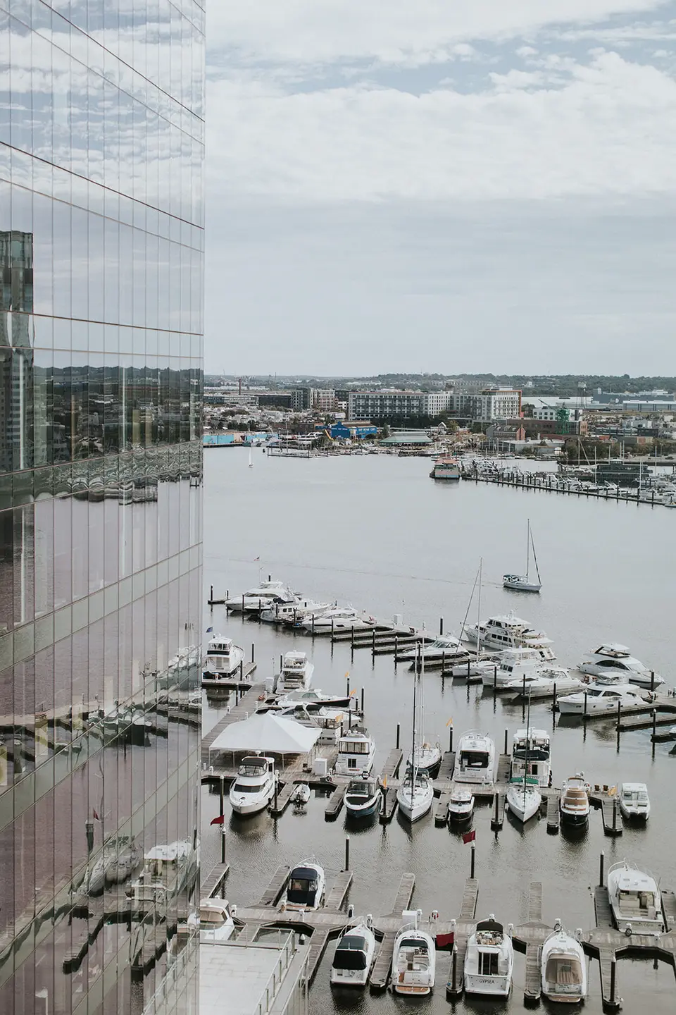 View of boats docked in a harbor beside a glass-walled building under a cloudy sky