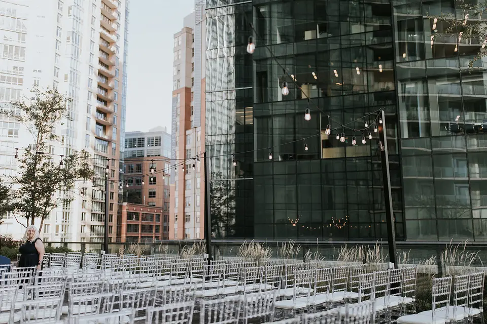 Rows of white chairs set up for an outdoor rooftop ceremony beside glass high-rise buildings