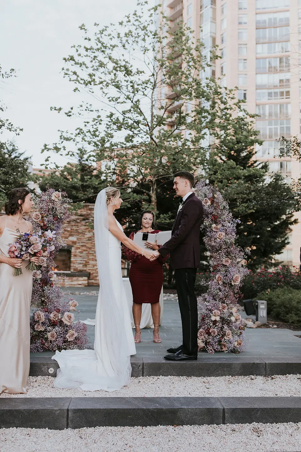 Bride and groom holding hands at a rooftop ceremony between floral arrangements with an officiant and a bridesmaid