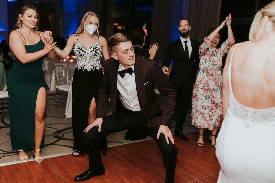 Groom in a burgundy tuxedo jacket crouching low on the dance floor as guests dance and clap around him