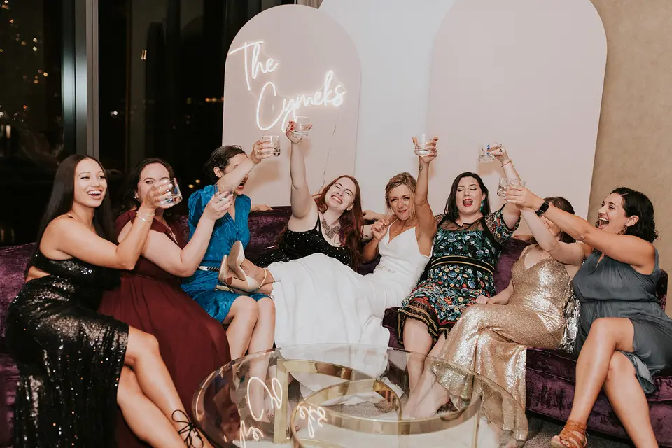 Bride and guests in formal dresses raising glasses on a purple velvet sofa beside a neon wedding sign