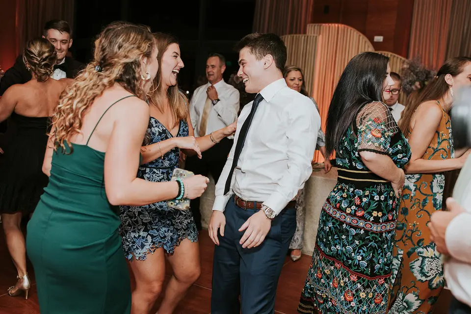 Guests dancing and laughing together on a wedding dance floor, a man in a white shirt and tie at center
