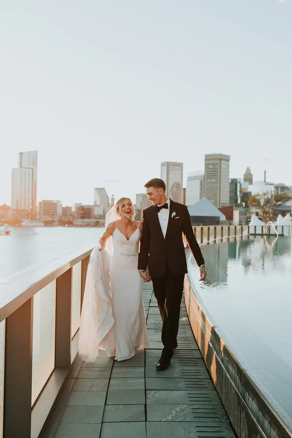Bride and groom walking hand in hand along a waterfront deck with a city skyline behind them