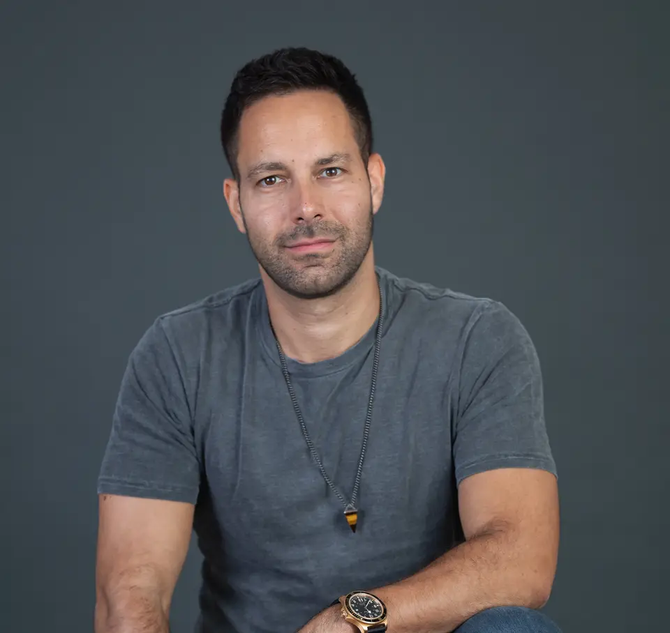 Man with dark hair and a gray t-shirt and pendant necklace posing against a dark gray backdrop