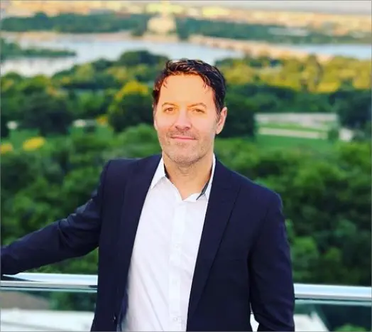 Man in a navy blazer and white shirt smiling on a rooftop with trees and a river behind