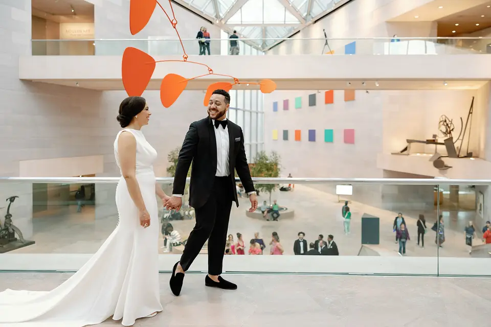 Bride and groom in a bow tie hold hands in a bright museum atrium with colorful art and an orange mobile above