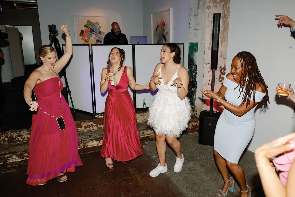 Four women in pink, red, white and light blue dresses dancing at a wedding reception with the DJ behind a white booth