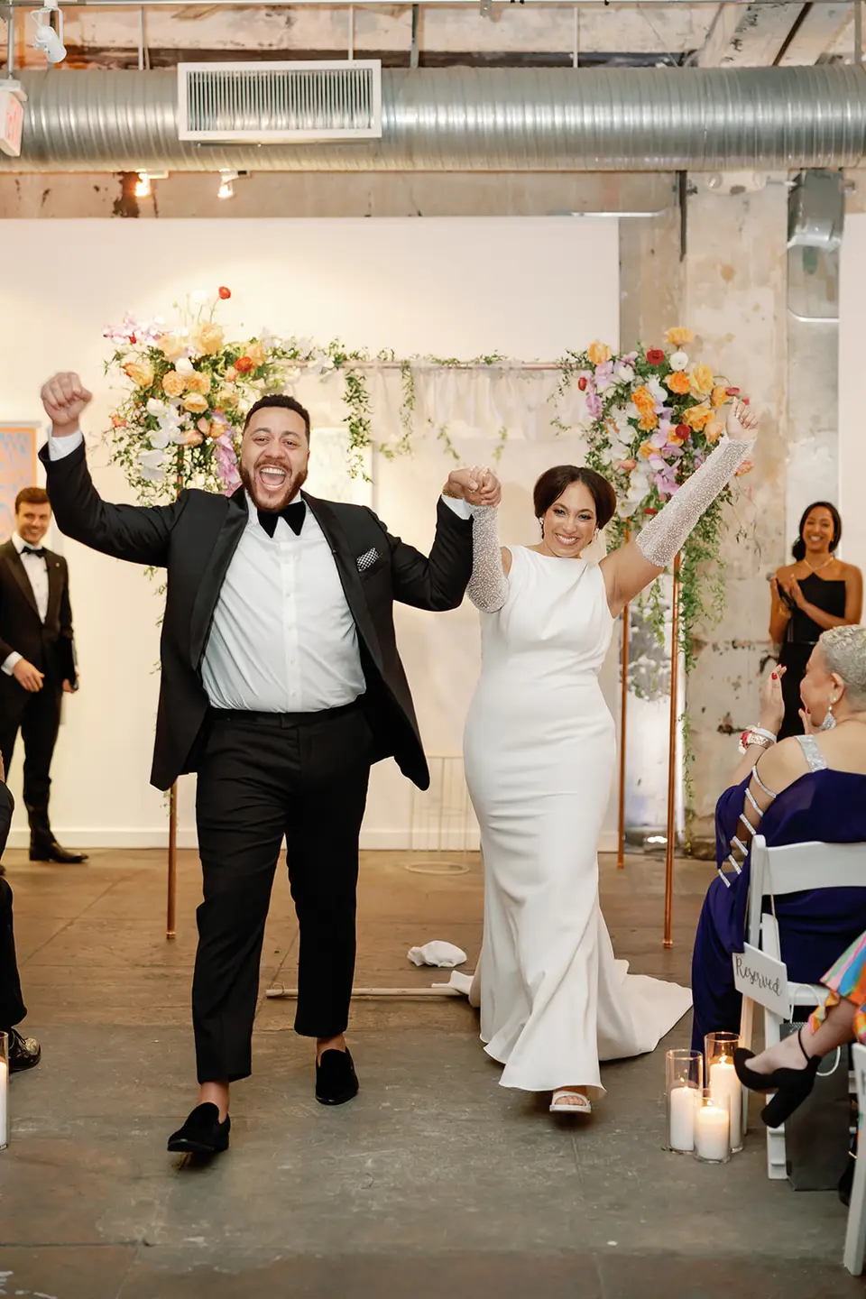Bride and groom raise their fists and smile as they walk up the aisle in front of a flower-covered arch in a loft space