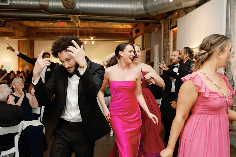Groom in tuxedo and bridesmaids in pink dresses dancing together on a wedding dance floor in an art gallery space