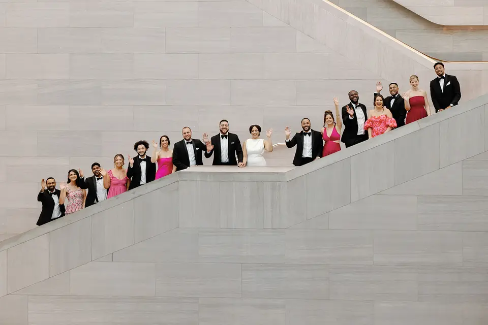 Bride, groom and wedding party in formal wear wave from a wide stone staircase inside a museum