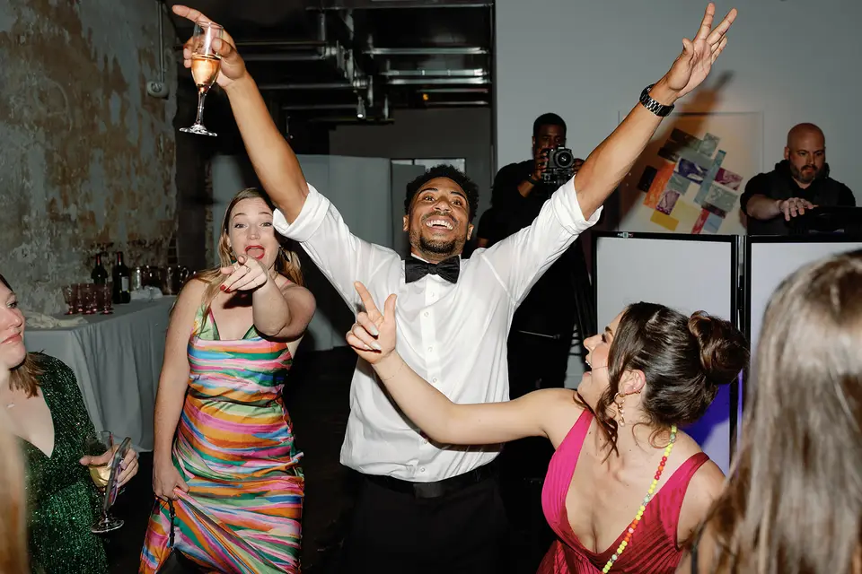 Groom in a bow tie with both arms raised dancing with guests as a photographer and DJ look on in the background