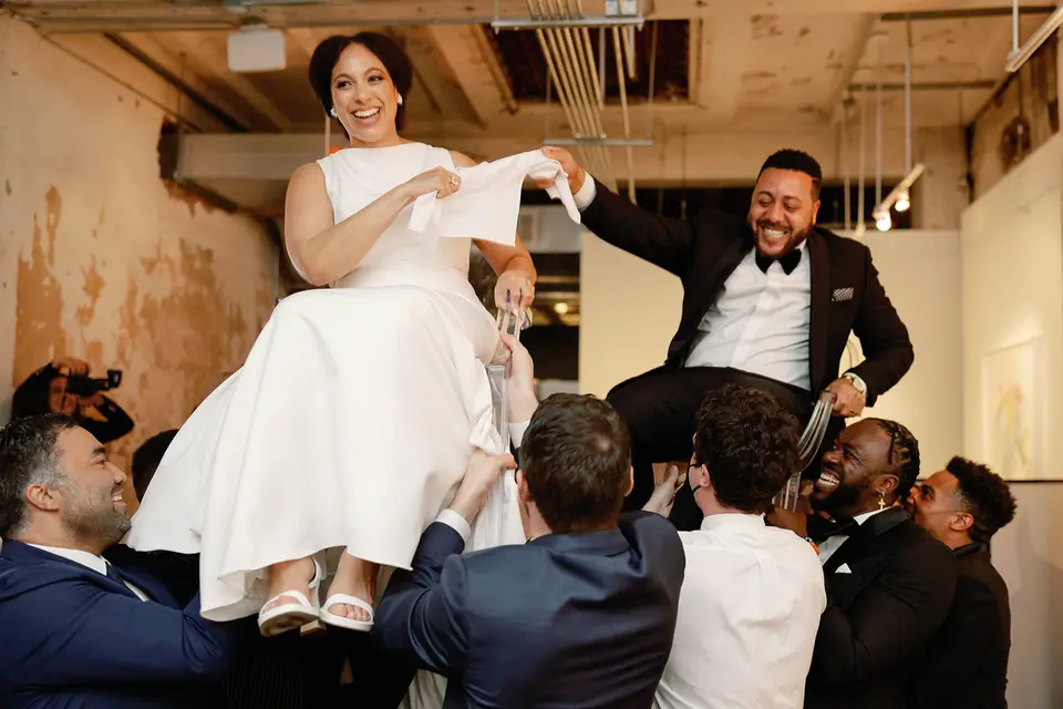 Bride and groom lifted in chairs by guests during a hora dance at a wedding reception in a gallery venue