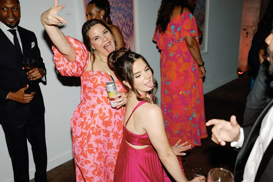 Two women in pink dresses dancing and sticking out their tongues at a wedding reception, one holding a drink