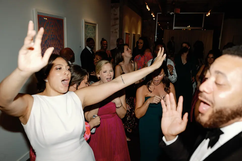Bride in a white dress with an arm raised dancing alongside a groom in a bow tie and guests at the reception