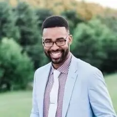 Smiling bearded man with glasses in a light blue blazer standing outdoors with trees behind