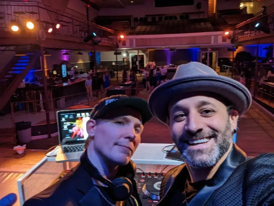 Two DJs taking a selfie behind a laptop and mixer in a large venue with purple uplighting