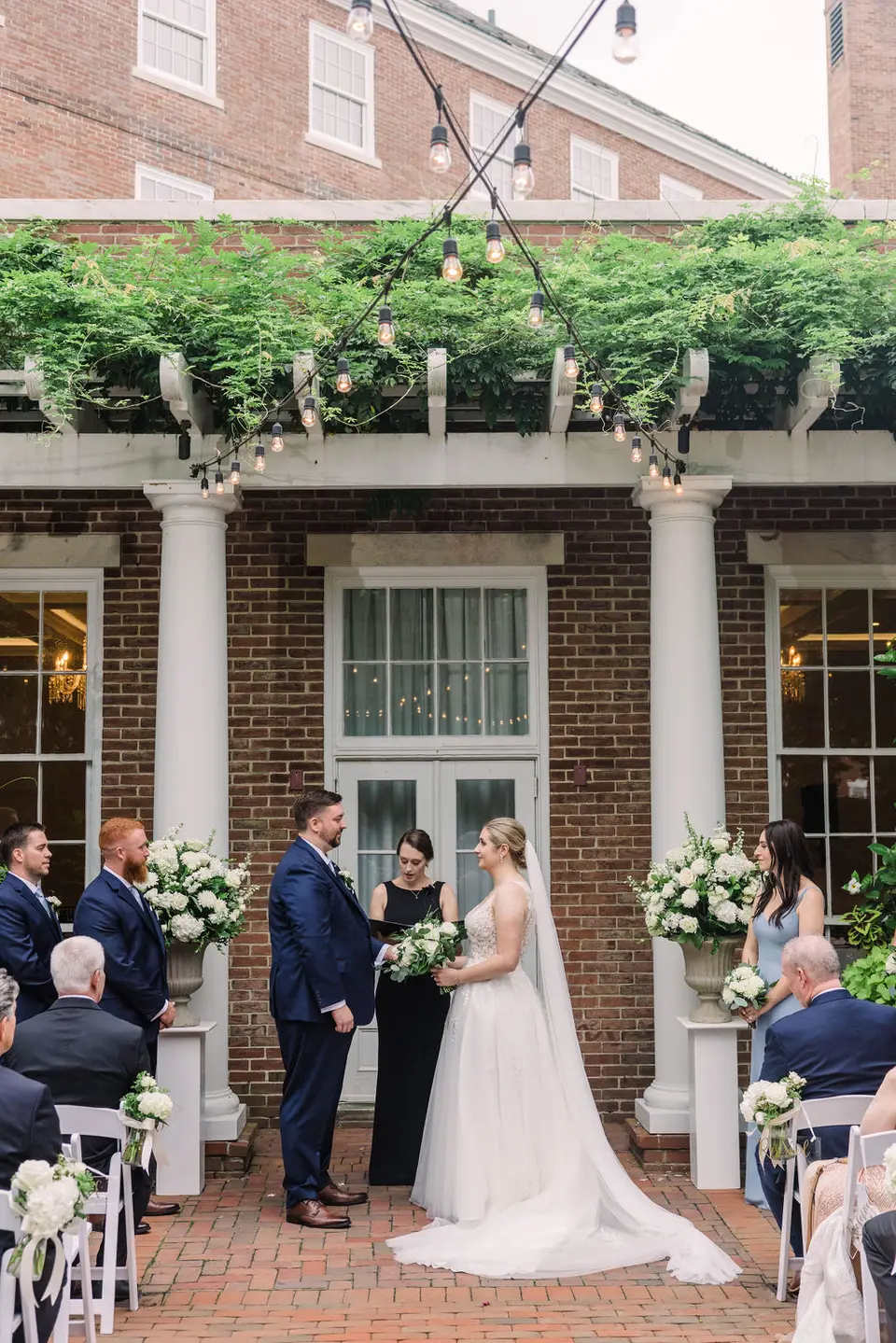 Couple facing the officiant at an outdoor brick courtyard ceremony with flowers and seated guests