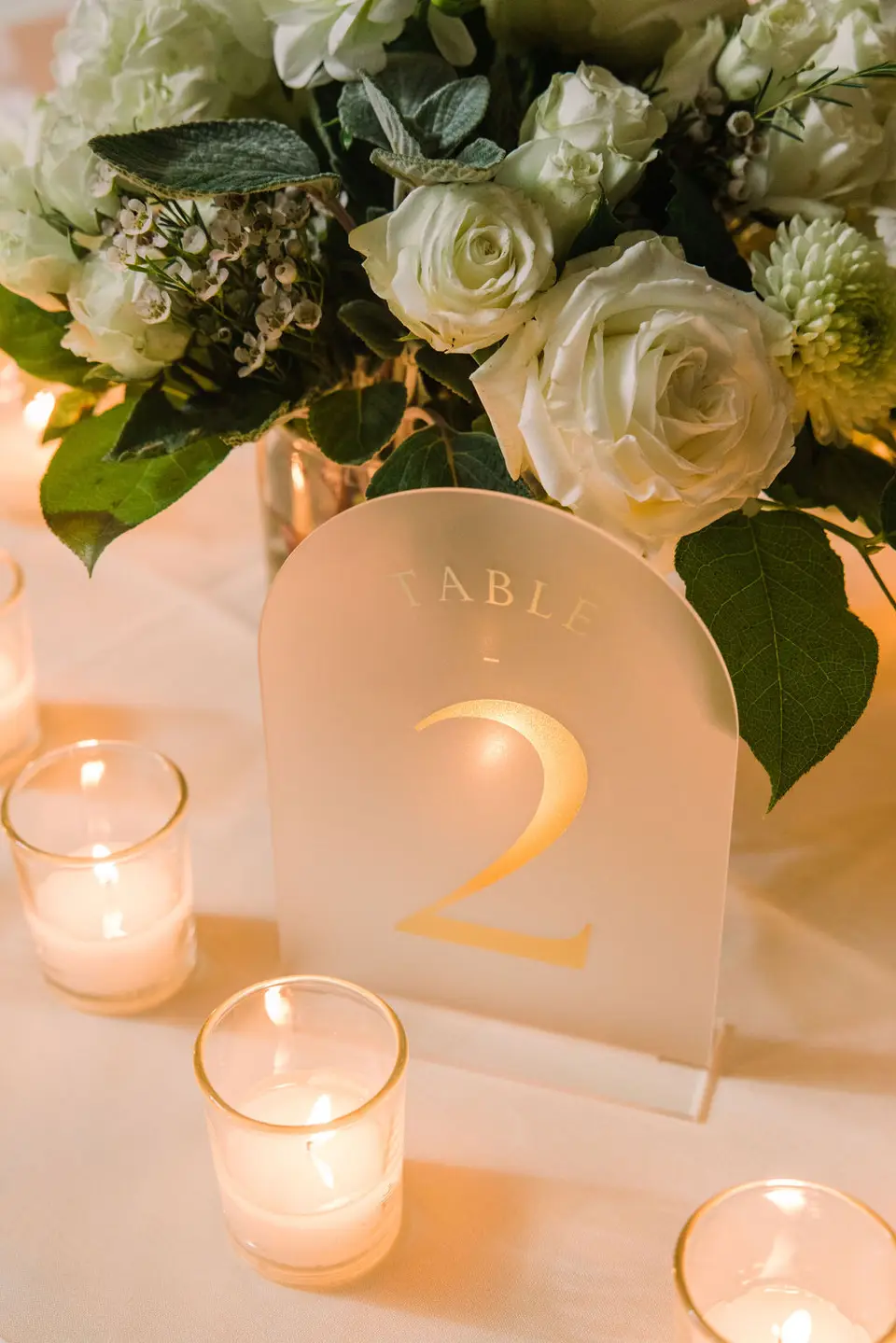 Frosted acrylic table number 2 sign beside white roses and glass votive candles