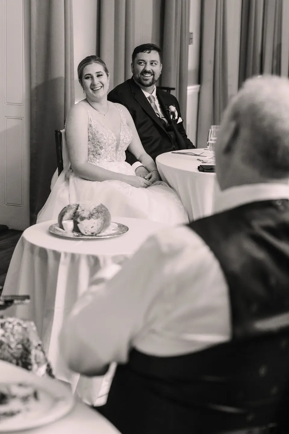 Black and white photo of the bride and groom smiling at their table with an older guest in the foreground