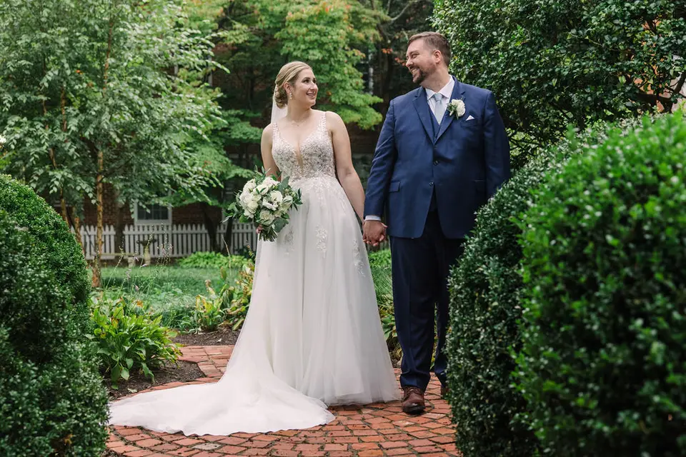 Bride and groom in a navy suit holding hands on a brick path in a green garden