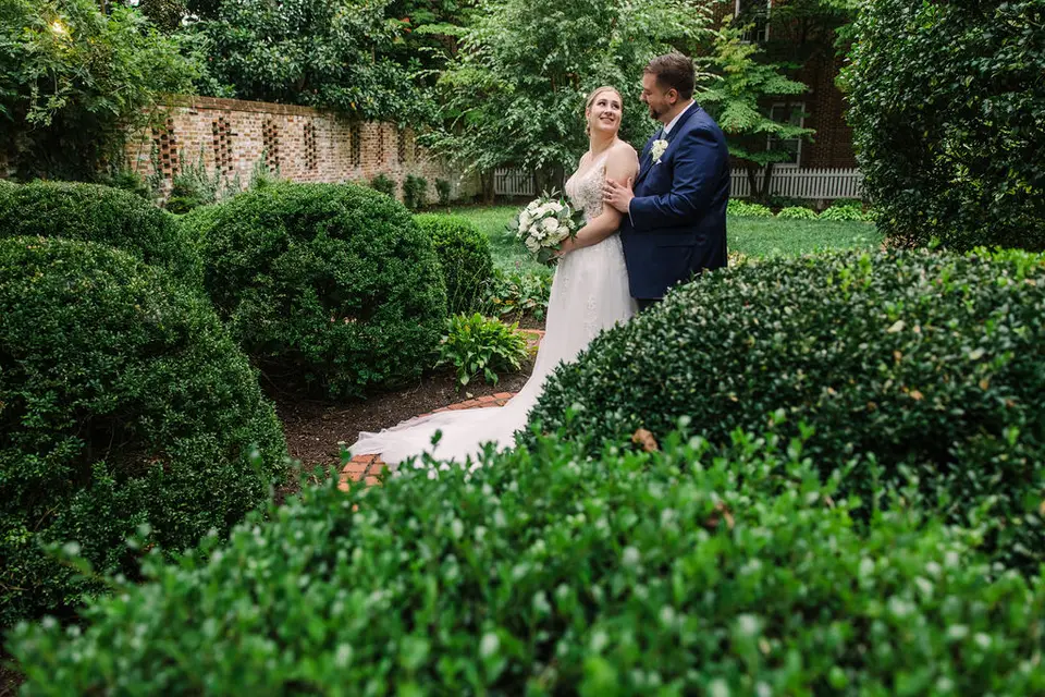 Bride holding a bouquet embraces the groom among trimmed boxwood hedges in a garden