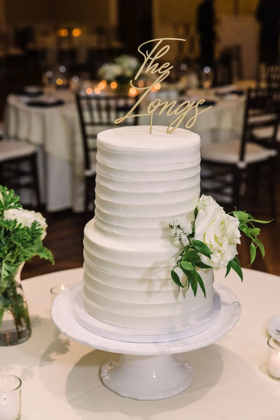 Two-tier white wedding cake with white roses and a gold topper reading The Longs