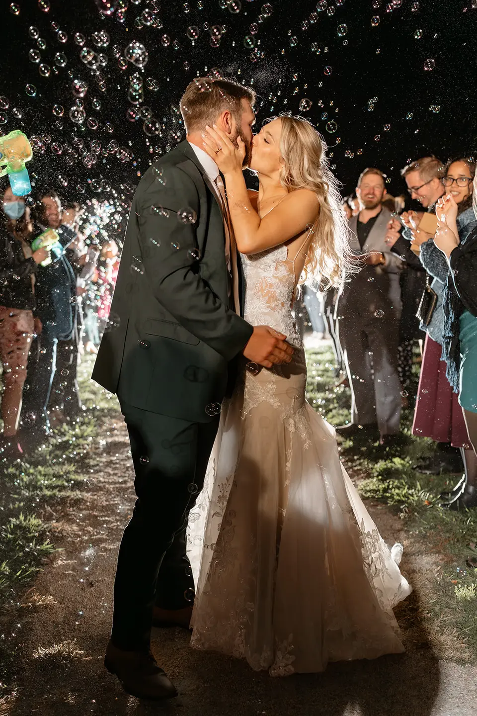 Bride and groom kissing as guests blow bubbles around them at night