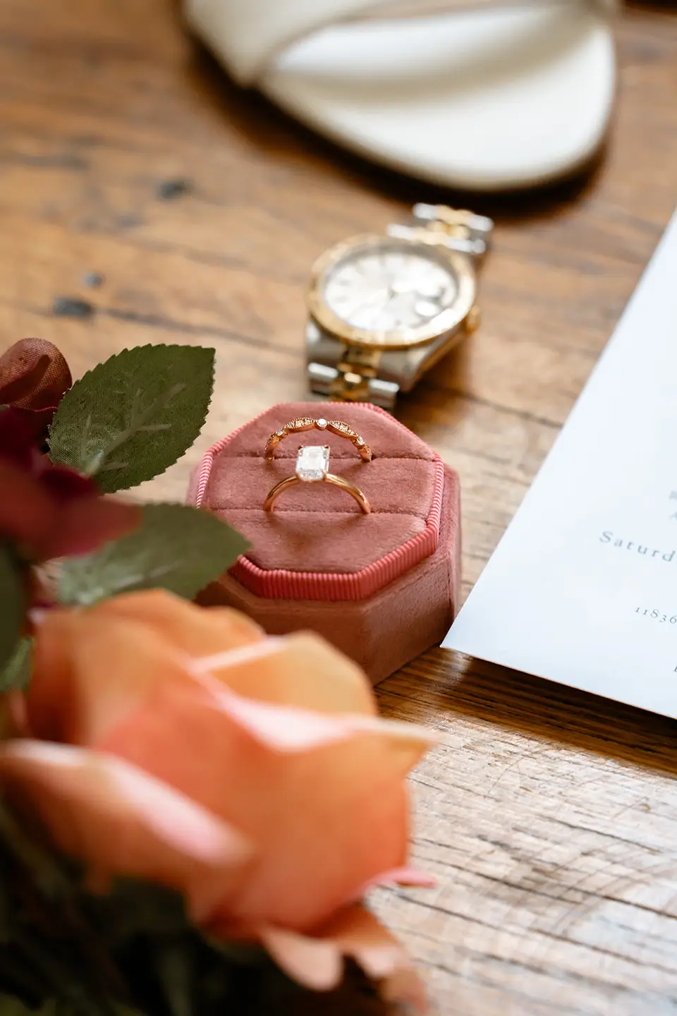 Engagement ring in a pink velvet box beside a wristwatch, white shoe, invitation and roses