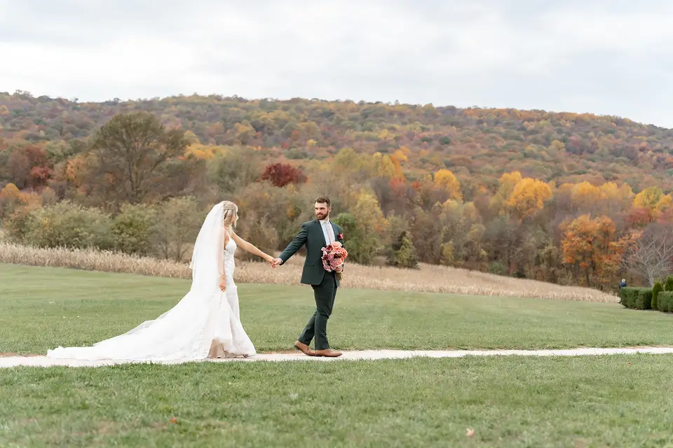 Bride and groom hand in hand walking across a lawn with autumn-colored hills behind them