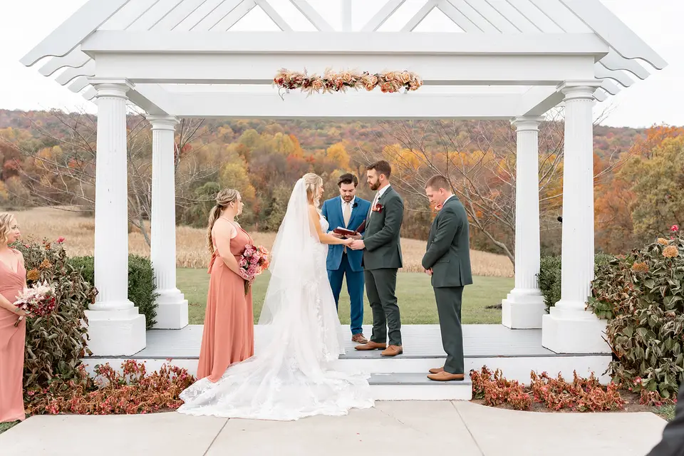 Couple exchanging vows under a white pavilion with autumn hills and bridesmaids in coral dresses