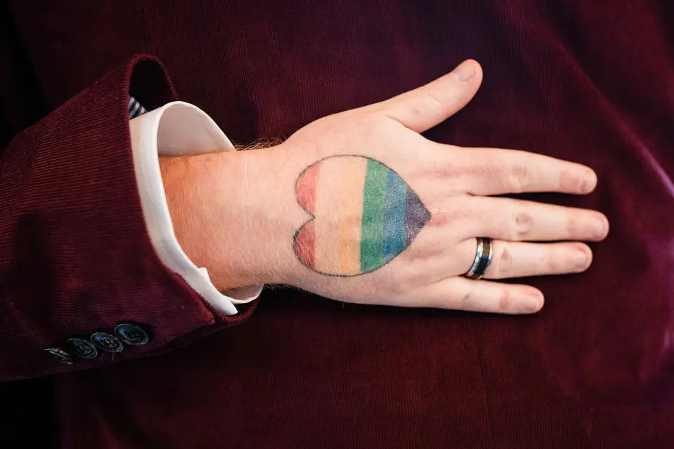 Close-up of a groom's hand with a rainbow heart drawn on it and a ring, resting on a burgundy jacket