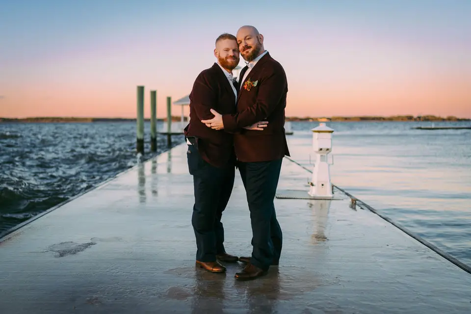 Two grooms in burgundy jackets embracing on a wooden pier at sunset