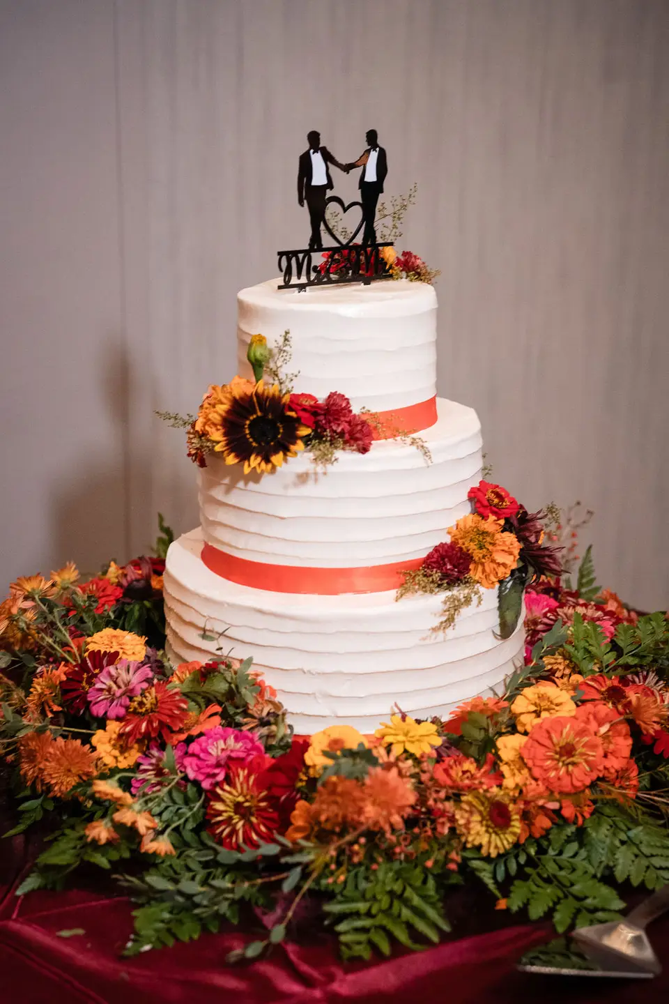 Three-tier white wedding cake with orange ribbon, fall flowers and a topper of two grooms
