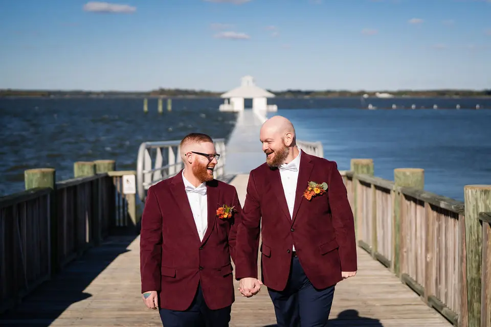 Two grooms in burgundy jackets holding hands and walking on a wooden dock by the water