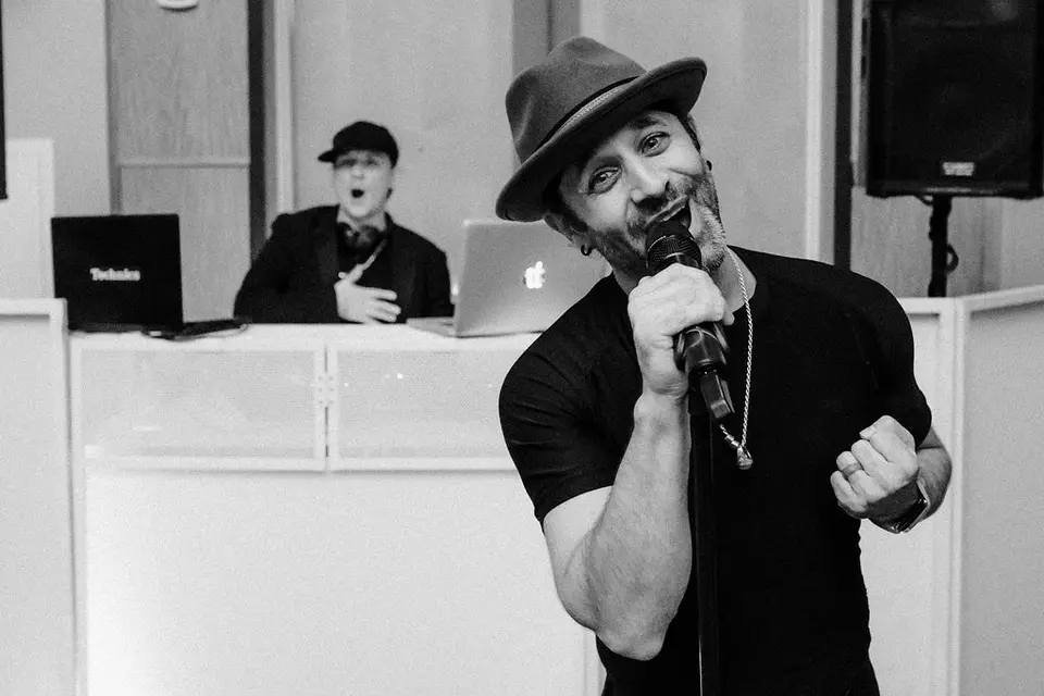 Man in a hat singing into a microphone in front of a DJ booth with a DJ behind it, black and white