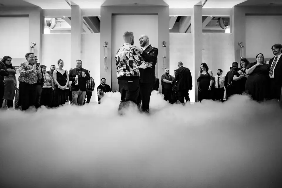 Two grooms dancing in low fog on a ballroom floor as guests watch, black and white