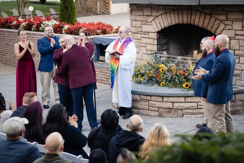 Two grooms hugging at an outdoor ceremony while an officiant in a rainbow stole and guests applaud