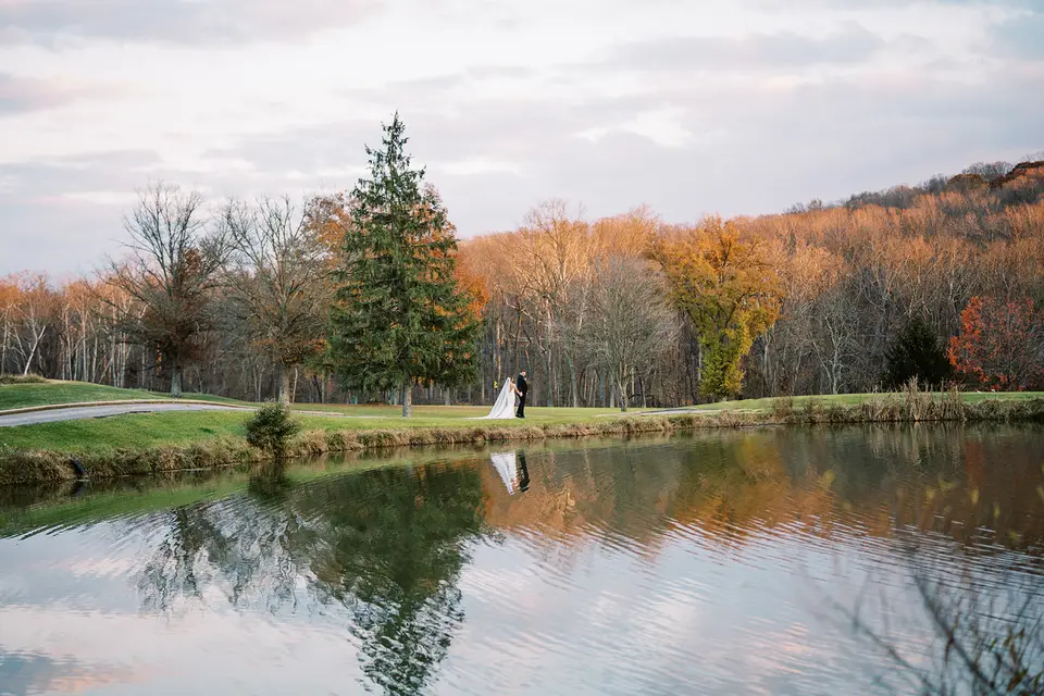Distant bride and groom stand by a pond reflecting autumn trees and a tall evergreen at sunset