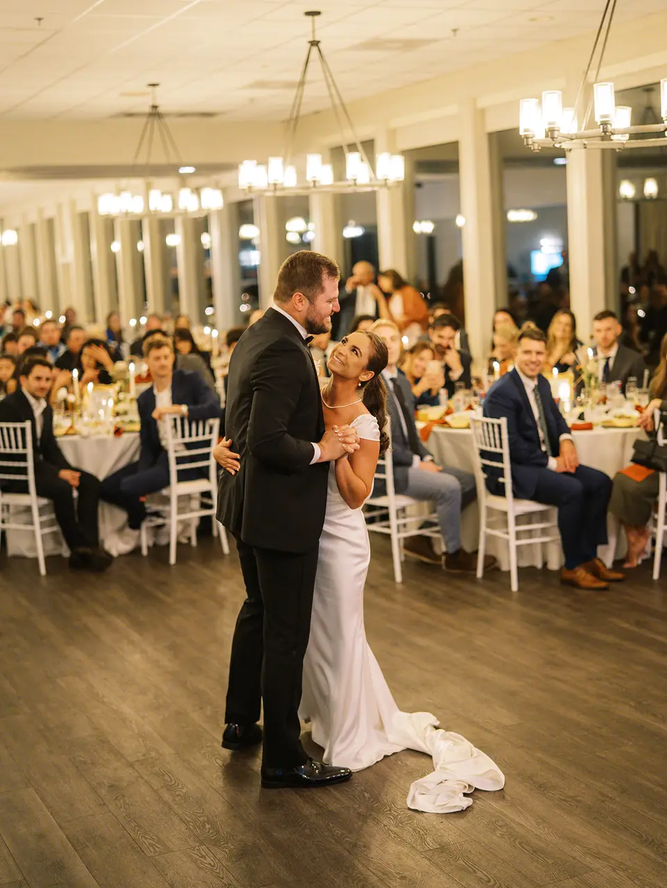 Bride and groom share a first dance on a wood floor as seated guests watch from round tables