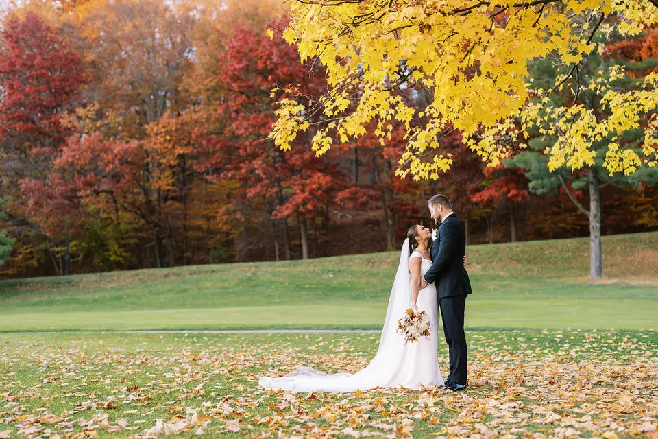 Bride and groom kiss on a golf course lawn under bright yellow and red autumn trees