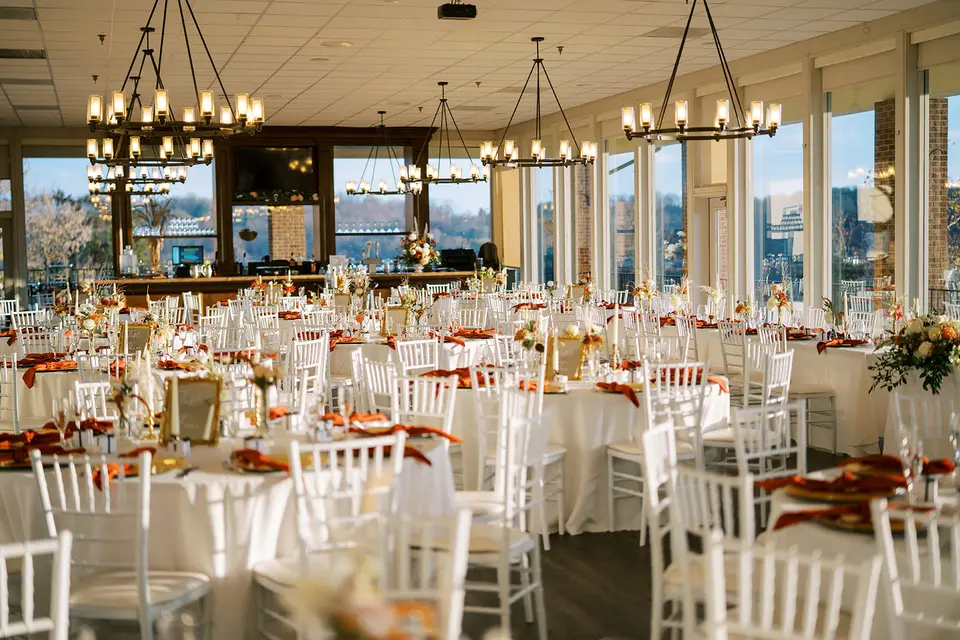 Reception room with white chairs, linen-covered tables, orange napkins, chandeliers and tall windows