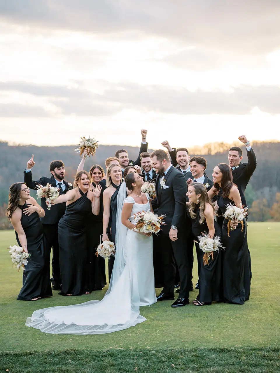 Bridal party in black cheer with fists raised around the bride and groom on a golf course at sunset