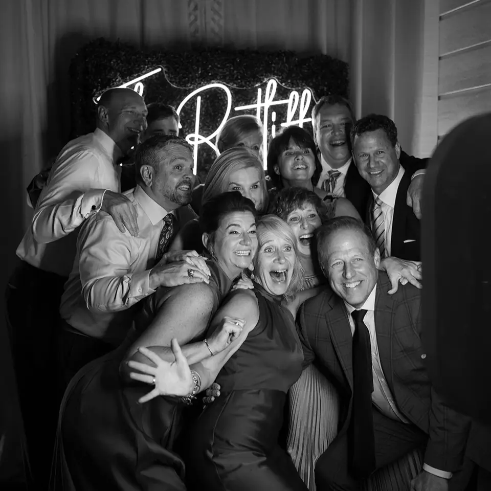 Black and white photo of a group of guests laughing together in front of a neon sign